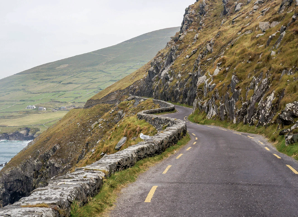 A winding Ireland coastal mountain road with a stone guardrail on the left, rocky hills on the right, and a view of green hills and farmland in the background.