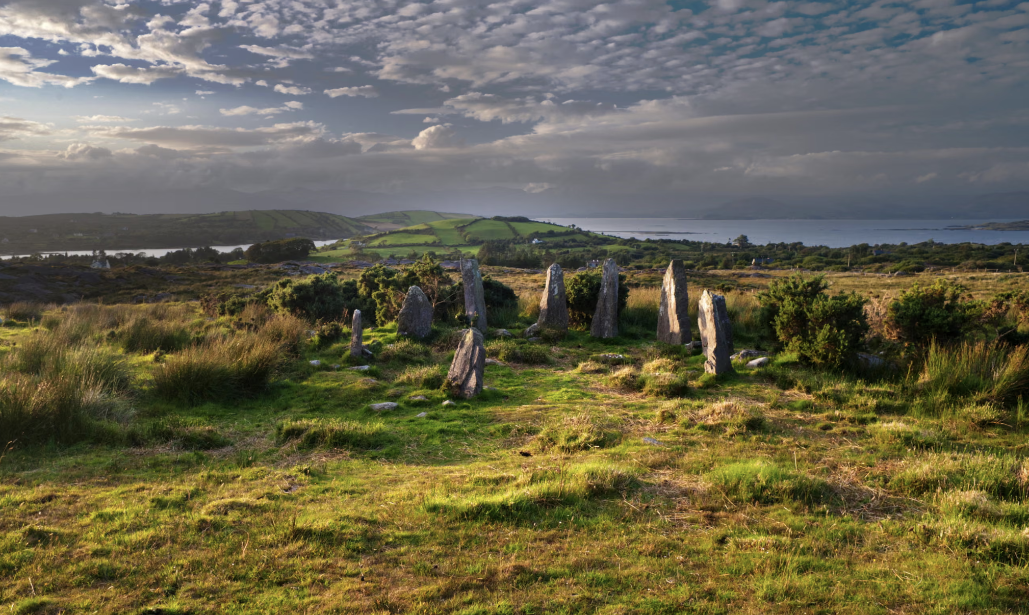 A scenic landscape featuring an ancient stone circle on a grassy hilltop with a body of water and rolling green hills in the background under a cloudy sky in south Ireland for a yoga retreat.