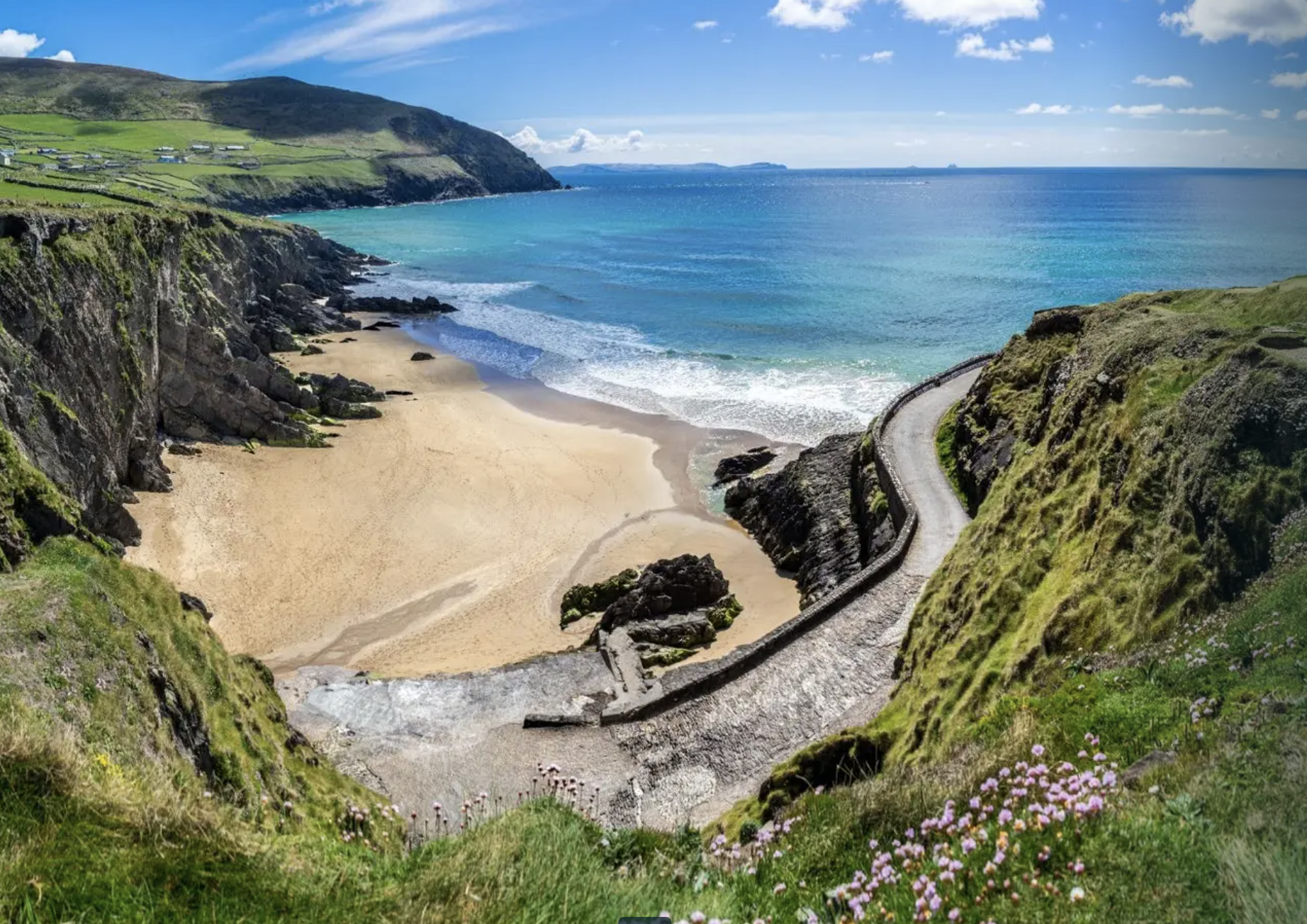 A coastal landscape with a sandy beach, rocky cliffs with green vegetation, a winding path, blue ocean waves, and a partly cloudy sky, taken from an elevated viewpoint in south Ireland.