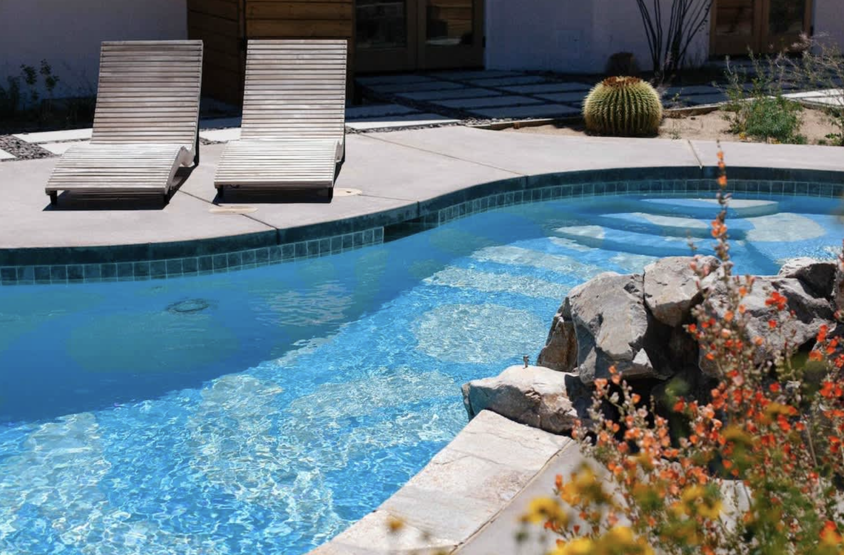A backyard swimming pool with two wooden lounge chairs on a concrete deck, desert plants including cacti, and a rocky water feature with orange flowers in the foreground.