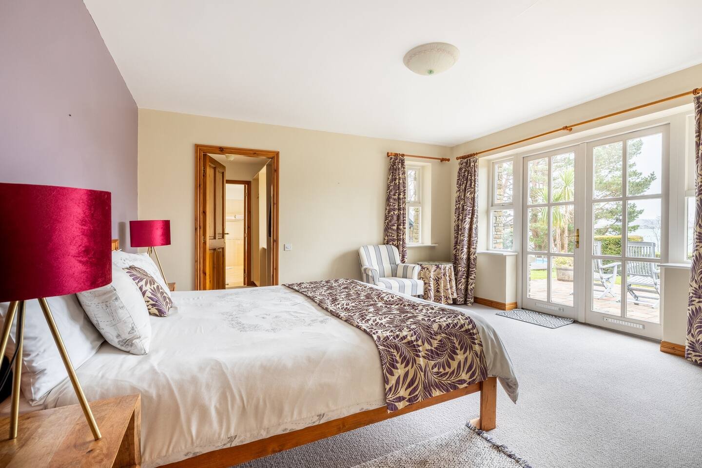Bright bedroom with large windows, bed with white bedding and floral-patterned blanket, striped armchair, two pink lamps, beige walls, wooden trim, and glass double doors leading to outdoor patio.