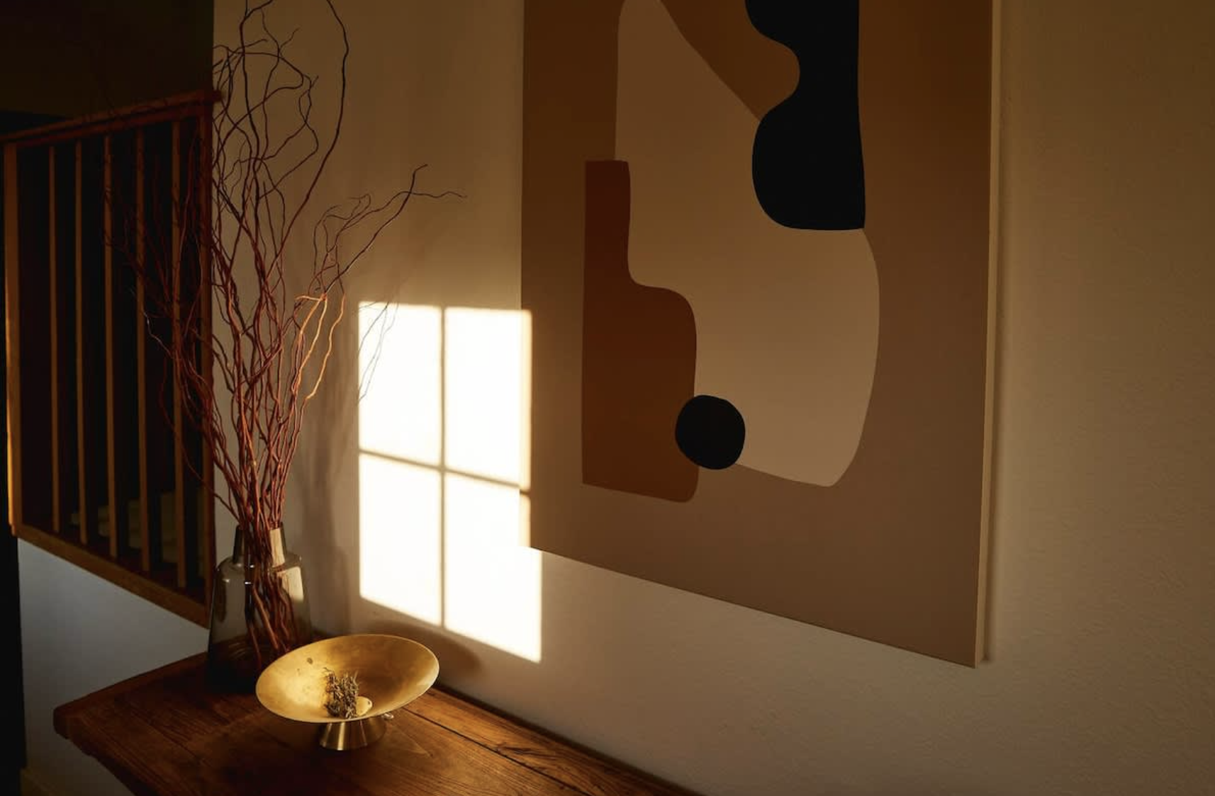 Sunlight casting a shadow of a window frame on a beige wall with a modern abstract painting. A wooden table holds a vase with red twigs and a gold decorative bowl.