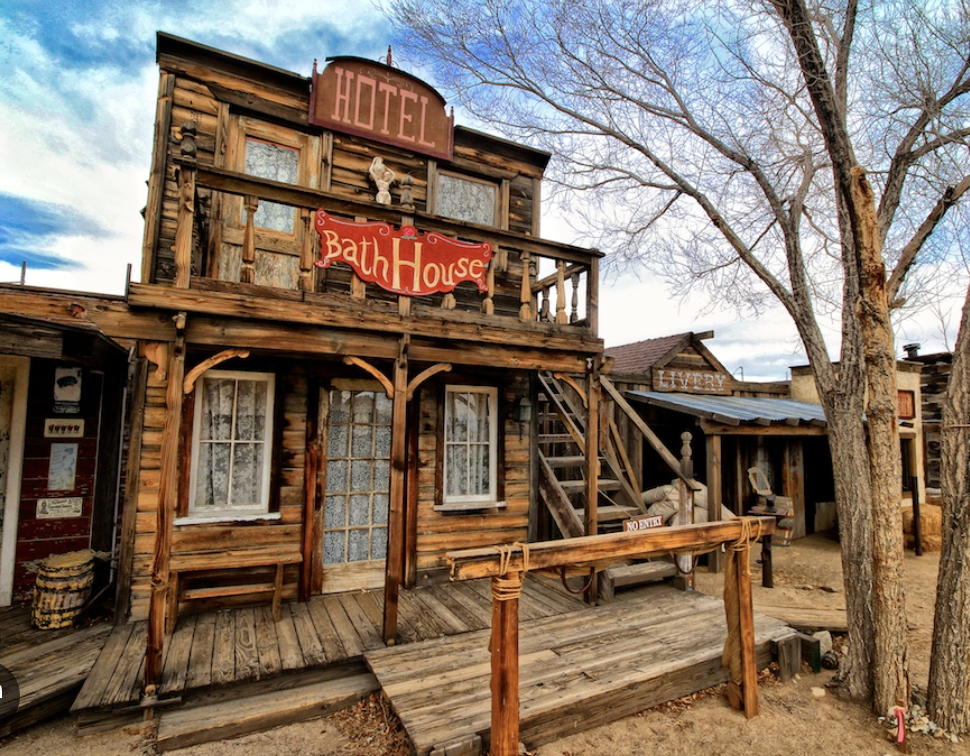 A rustic wooden building with a sign that reads 'Hotel' and another sign that says 'Bath House'; it has a balcony and stairs leading to the upper level, set next to a leafless tree on a dirt ground.