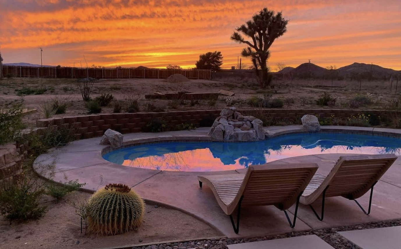 A desert backyard with a small kidney-shaped swimming pool reflecting a colorful sunset sky, with two wooden lounge chairs and desert plants including a large barrel cactus, and a tree silhouette in the distance.