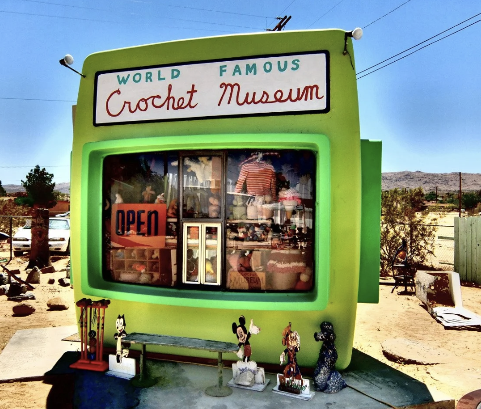 A small green building shaped like an old television set, with a sign that reads 'World Famous Crochet Museum.' The window display shows various crochet items, and there is a neon 'Open' sign. Decor items including cartoon character figures are placed in front of the building.