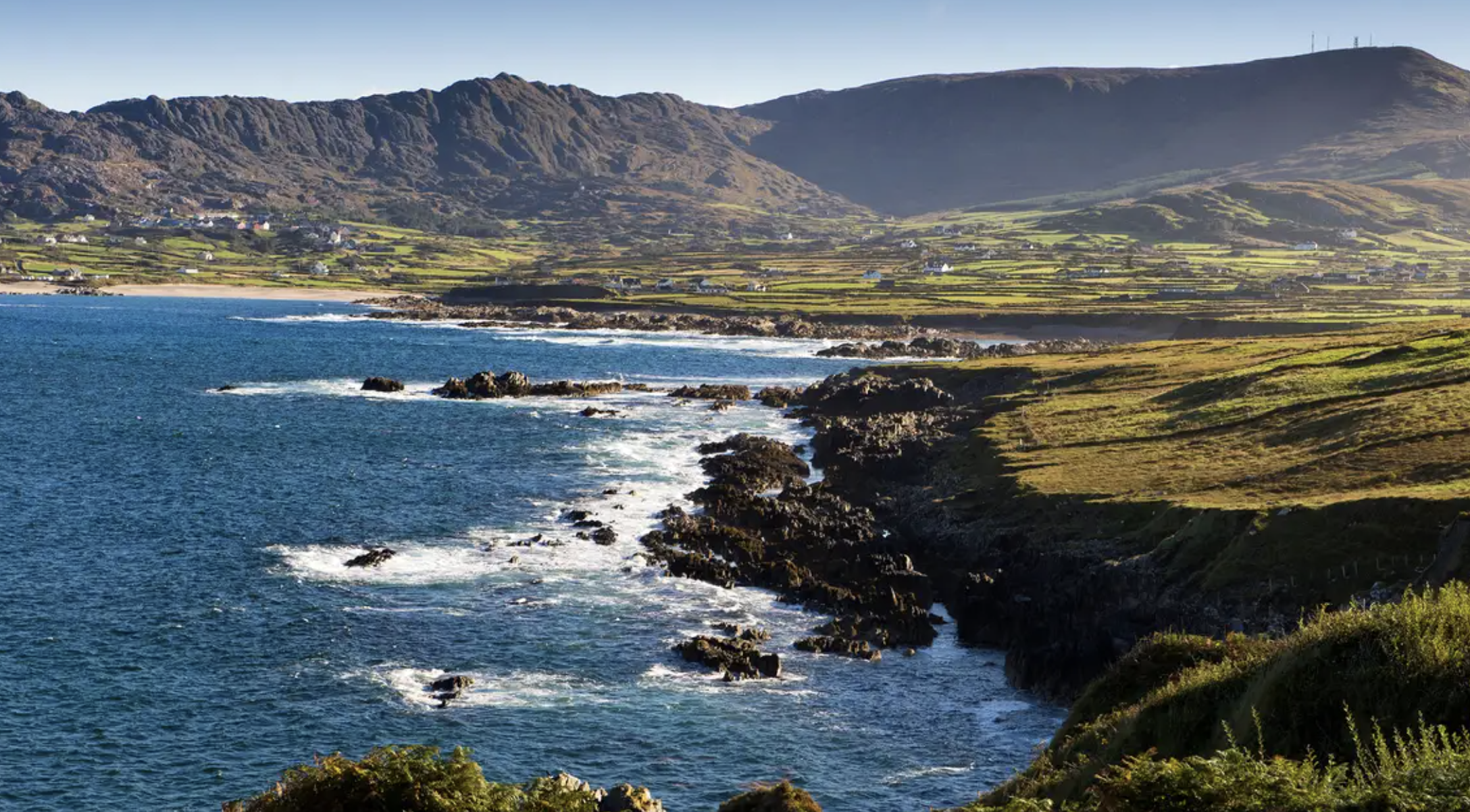 Scenic coastal landscape in Ireland with rocky shoreline, blue ocean waves, green fields, and mountain range in the background under clear sky.