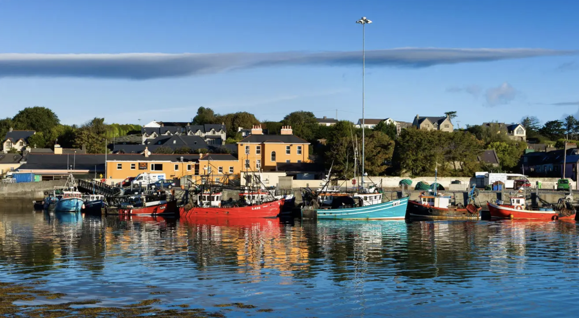 Boats docked in a harbor with colorful buildings and houses on a hillside in the background, under a blue sky with stretched cloud in south Ireland.
