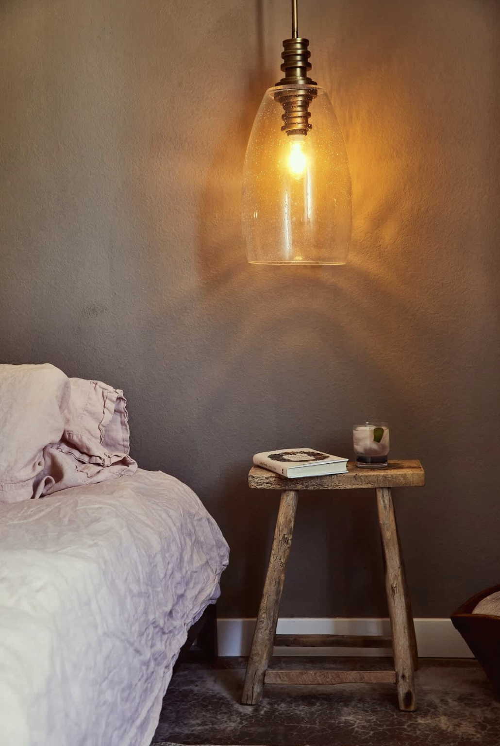 A cozy bedroom corner with a hanging glass pendant light, a bed with pink linens, a rustic wooden side table holding a book and a glass of drink on ice, and a textured wall background.