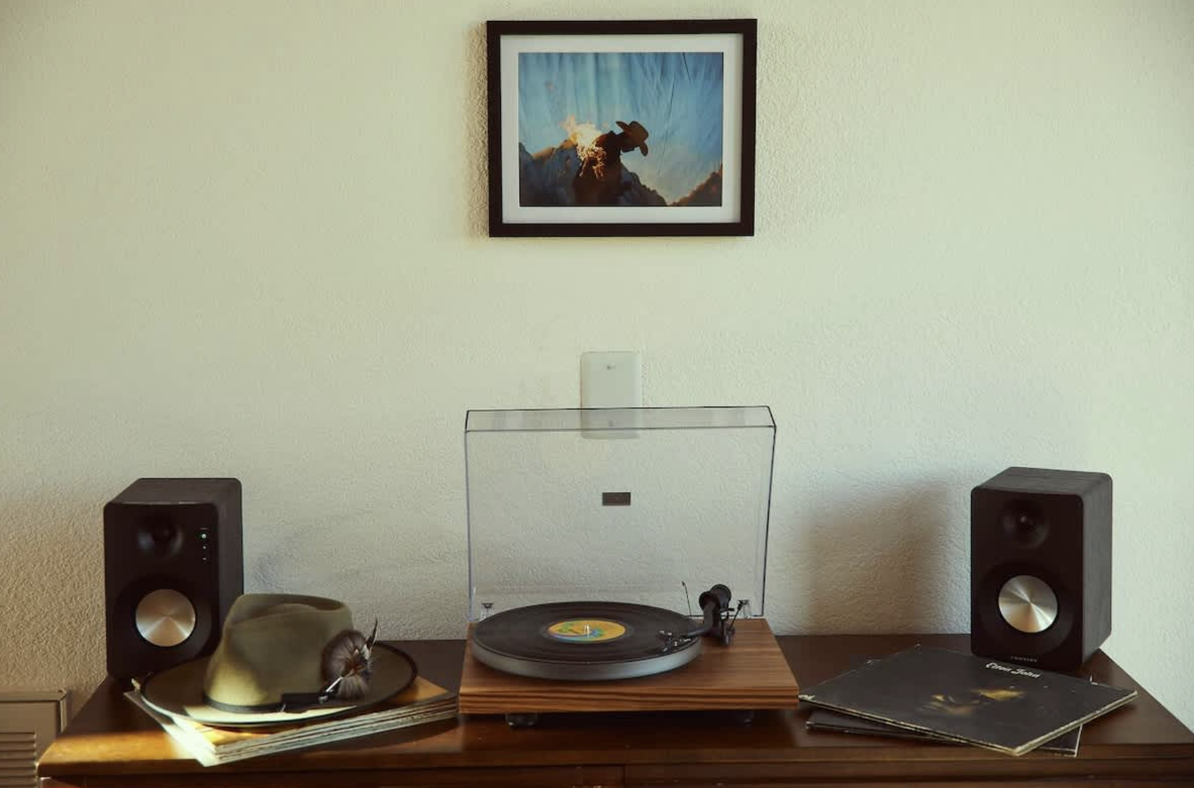 A turntable on a wooden table with speakers and vinyl records, positioned beneath a framed picture of a cowboy with a hat, against a plain white wall.