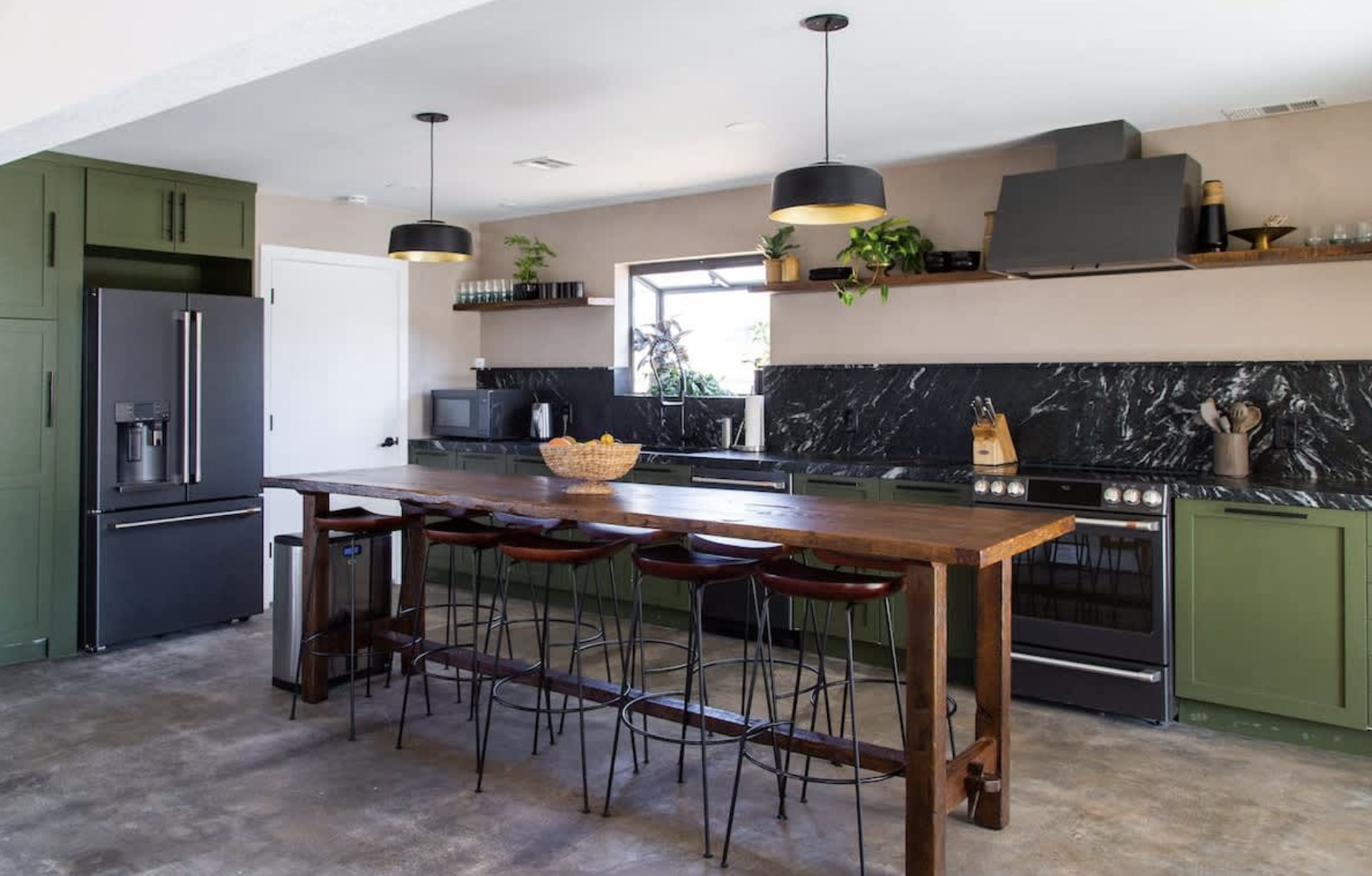 Modern kitchen with green cabinets, black marble backsplash, stainless steel appliances, a large wooden island with four bar stools, and open shelves with plants and kitchenware.