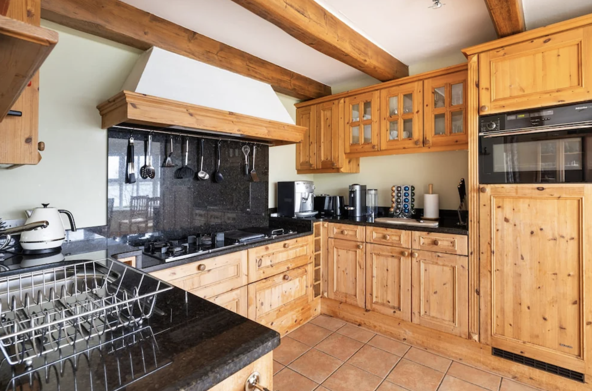 A kitchen with wooden cabinets, black countertops, a black backsplash, and a floor with brown tiles. There are various appliances including a coffee maker, a microwave, and a kettle. Cooking utensils hang above the stove at a yoga retreat.