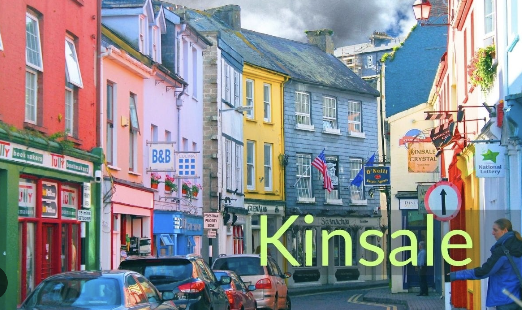 A colorful street scene in Kinsale, Ireland, for a yoga retreat, featuring brightly painted buildings in pink, blue, yellow, and red with parked cars along the street. There are signs for local businesses, flags, and a person walking on the sidewalk.