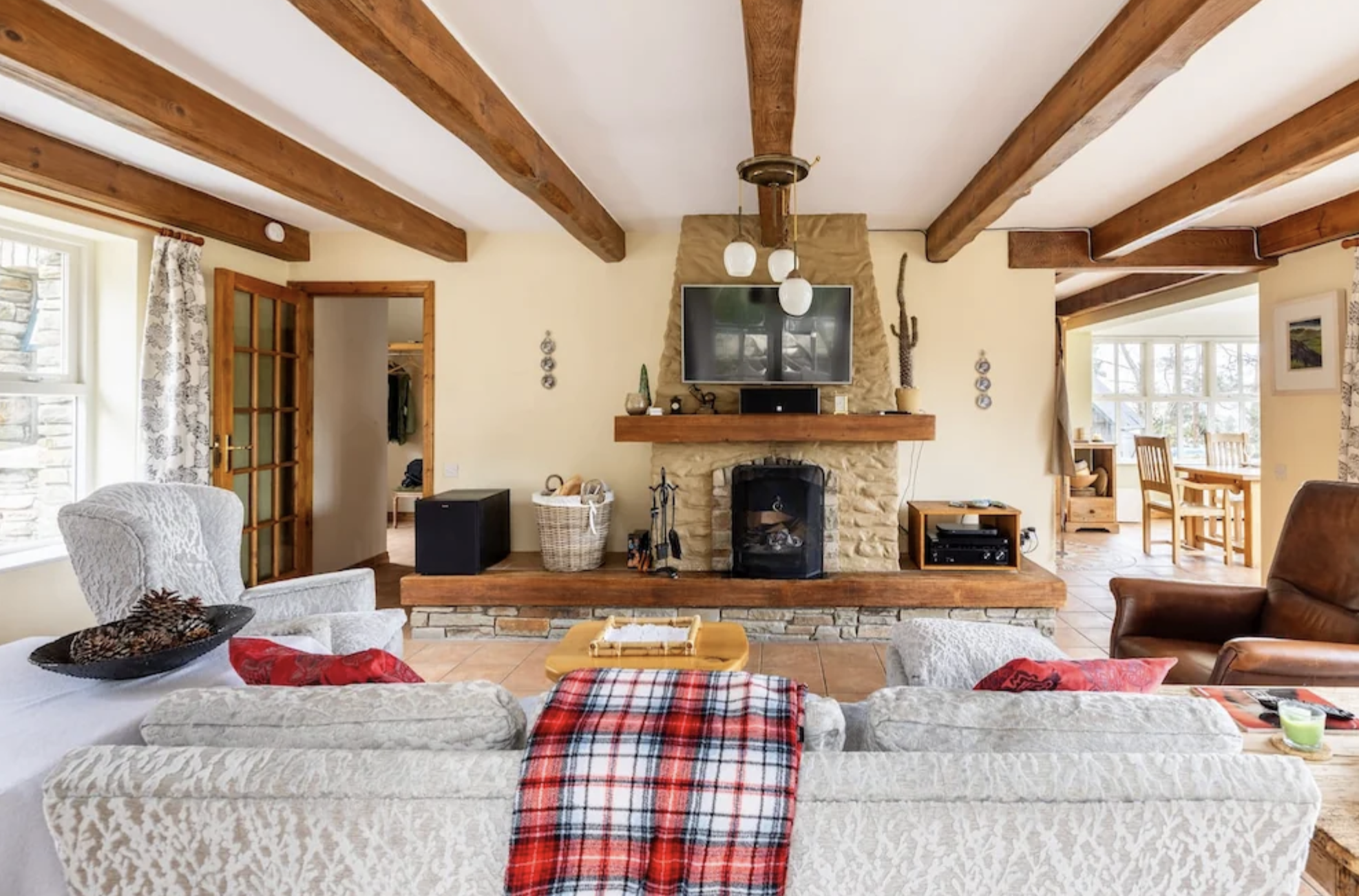 Retreat house in Ireland's cozy living room with a stone fireplace, wooden beams on the ceiling, a flat-screen TV above the fireplace, and comfortable armchairs and sofas. Decor includes a plaid throw, basket, and candles.