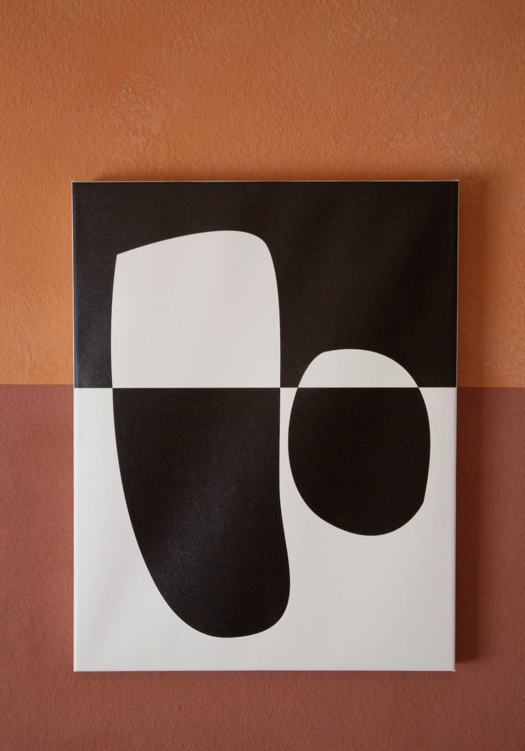 Abstract black and white artwork with organic shapes on a square canvas hanging on a two-tone orange and terracotta wall.