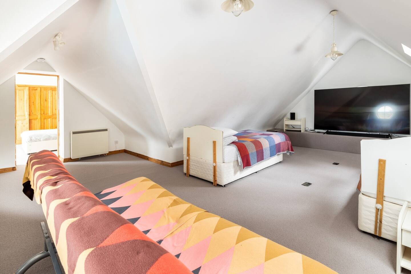 Spacious attic room with sloped ceiling, containing a television, a sofa, and beds, illuminated by natural light from windows.