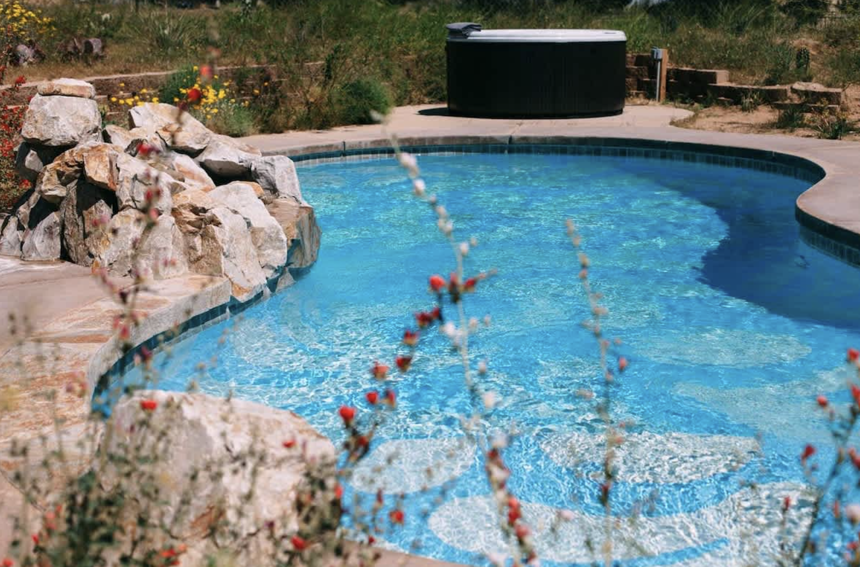 A backyard swimming pool with clear blue water, surrounded by stone and concrete pavements. There is a rock waterfall feature on the left, with plants and flowers around it. In the background, an outdoor hot tub is visible on a small concrete patio, with a grassy and slightly overgrown yard beyond.