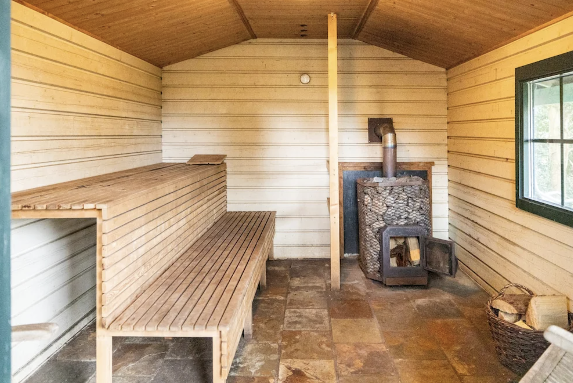 Sauna at a yoga retreat with wooden walls, wooden bench seating, a cast iron stove, and a window.