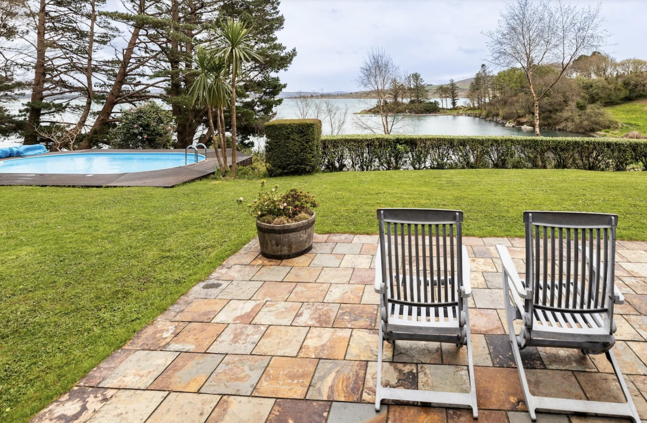 Retreat house in Ireland's backyard patio with two lounge chairs facing a grassy lawn and a lake, surrounded by trees and shrubs, with a hot tub on the left side.