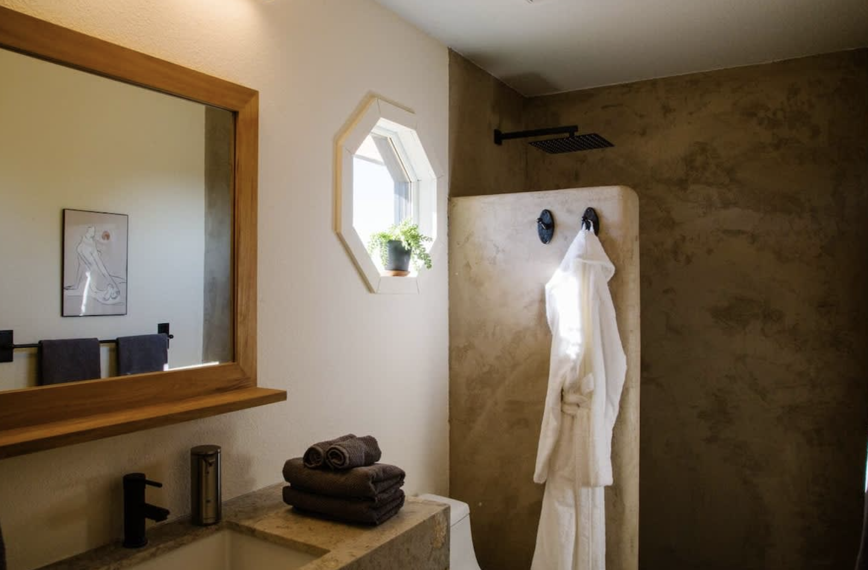 Modern bathroom with a large mirror, a small window, and a shower area with a black rain showerhead. A white bathrobe hangs on a hook, and a stack of folded towels sits on a countertop.