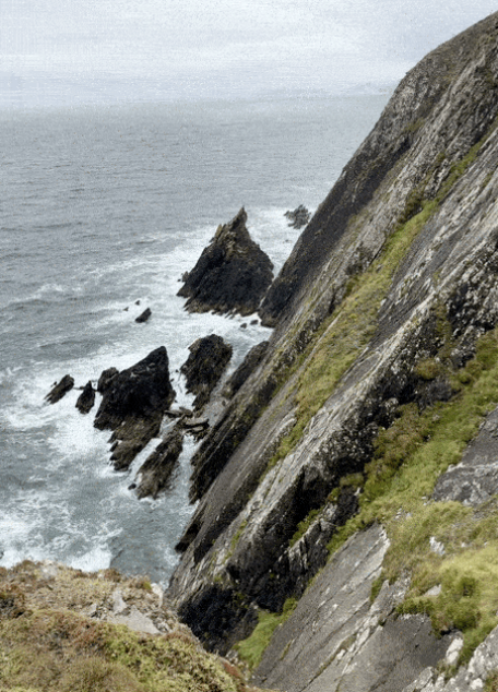 Cliffside overlooking rocky shoreline and ocean with waves, under cloudy sky.