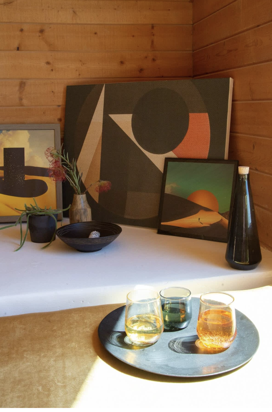 A tray with three glasses of colored drinks on a table in front of a wooden wall, with framed abstract and landscape art, a black vase, a small plant, and a bowl on a white surface behind.