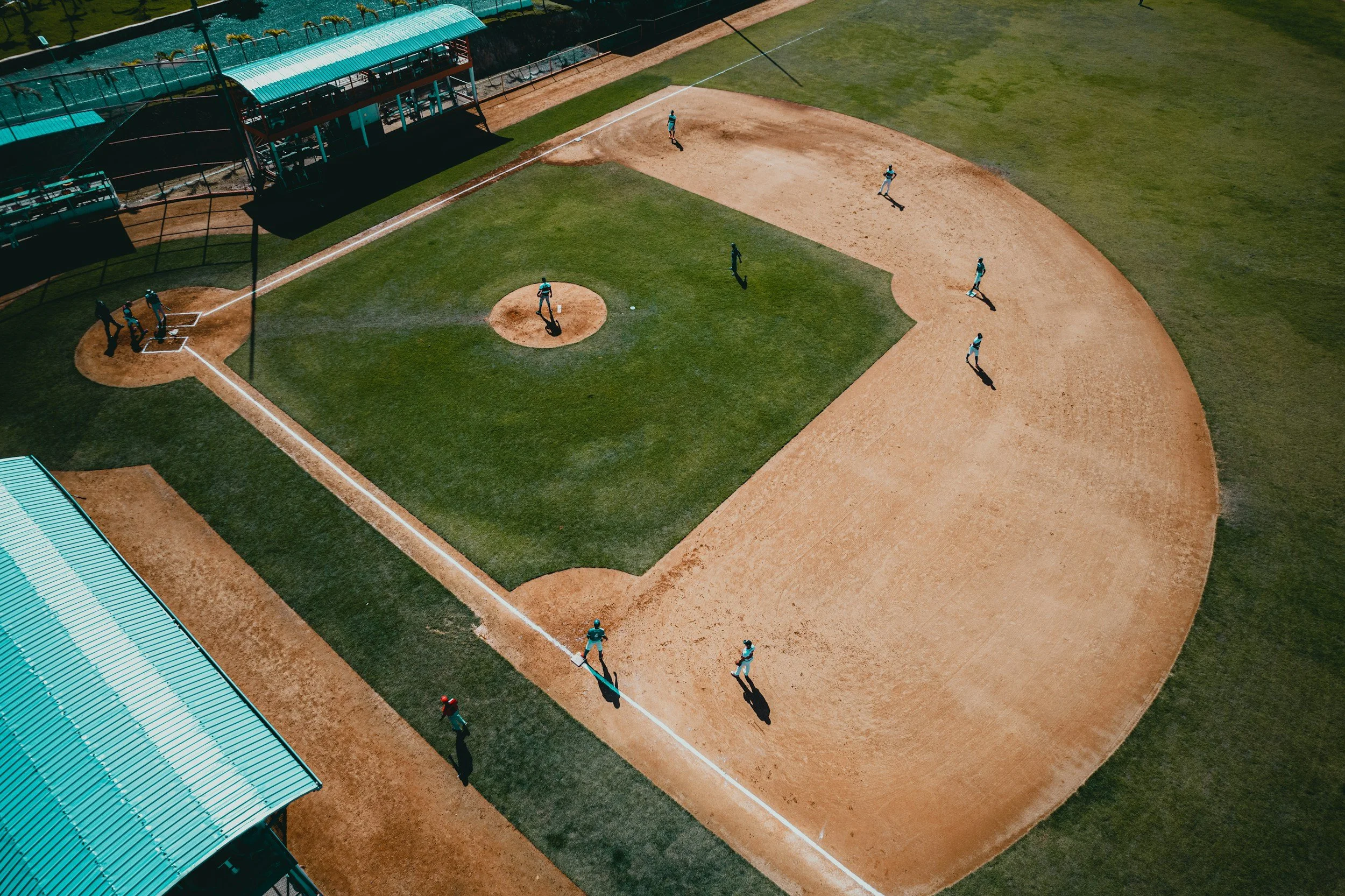 Aerial view of a baseball field with players practicing, including fielders and a pitcher, with team members near the dugout and shadows cast on the field from sunny weather.
