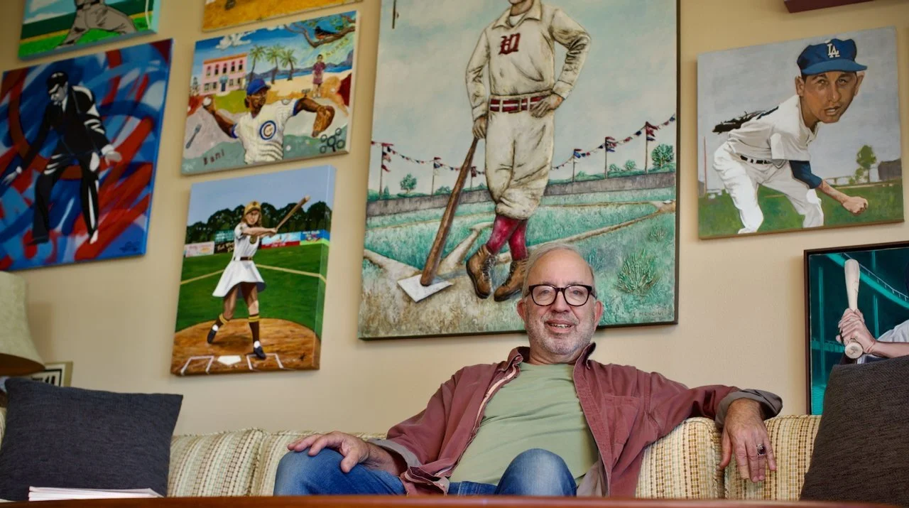 A man with glasses and a beard sitting on a sofa in a room decorated with vintage sports paintings, including baseball and beach scenes.