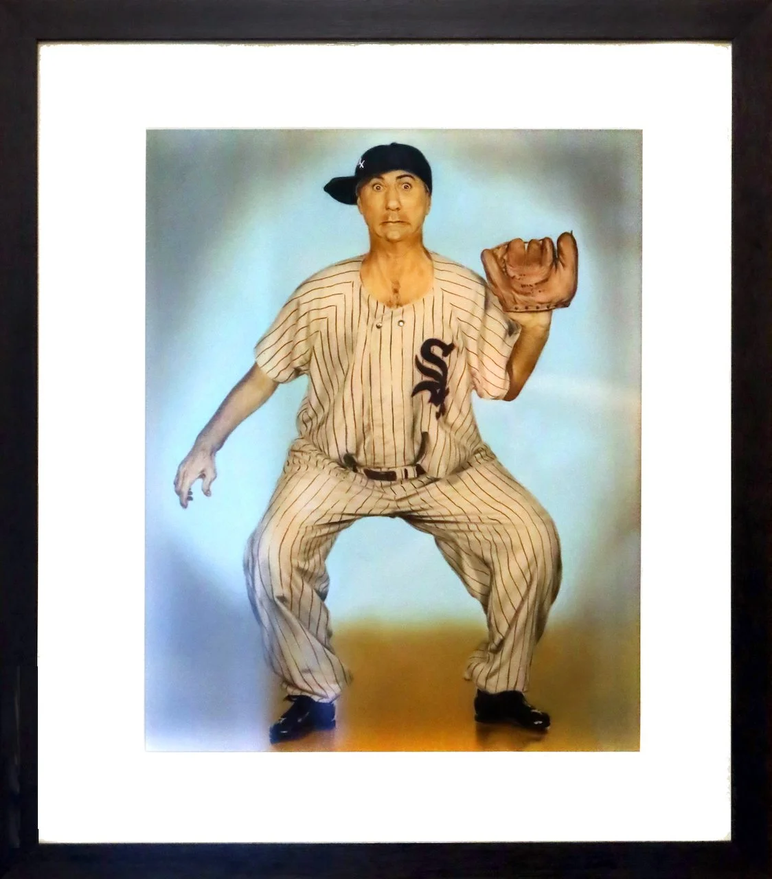 A person in a baseball uniform with a cap worn sideways, standing in a crouched position, holding a baseball glove, with a surprised expression on their face.