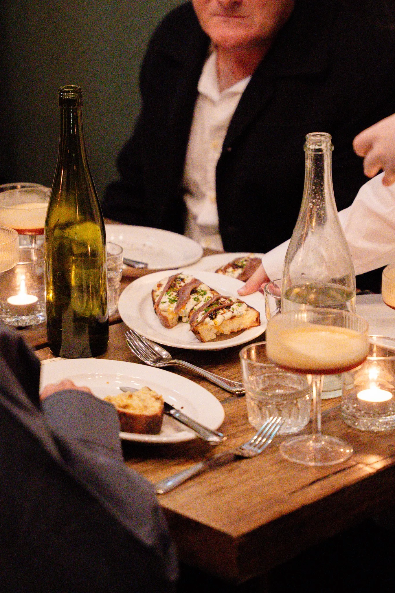 People dining at a restaurant with plates of food, glasses of wine, and lit candles on a wooden table.