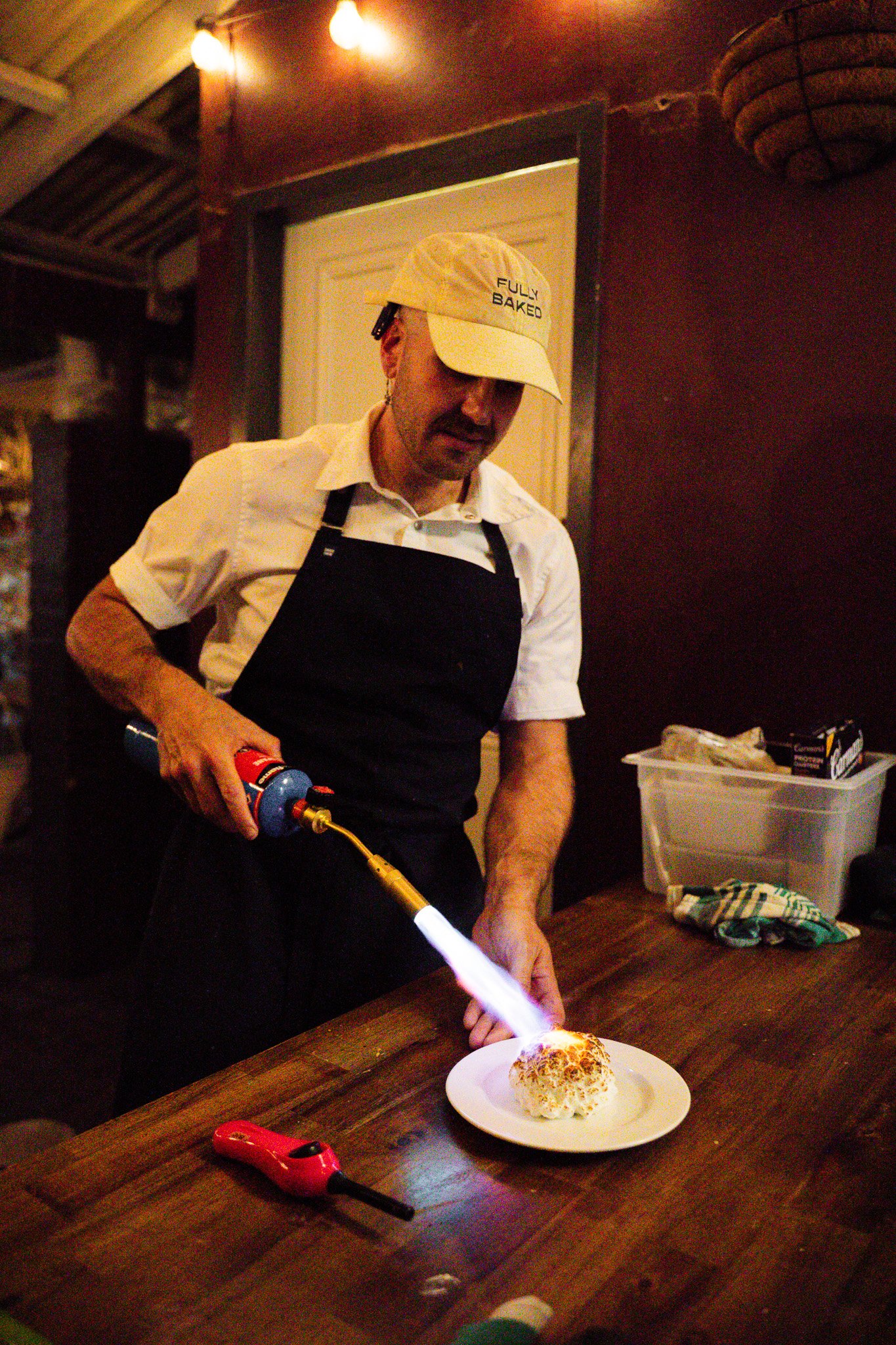 A man wearing a beige cap, white shirt, and black apron is torching a baked dish with a culinary blowtorch in a warmly lit kitchen.