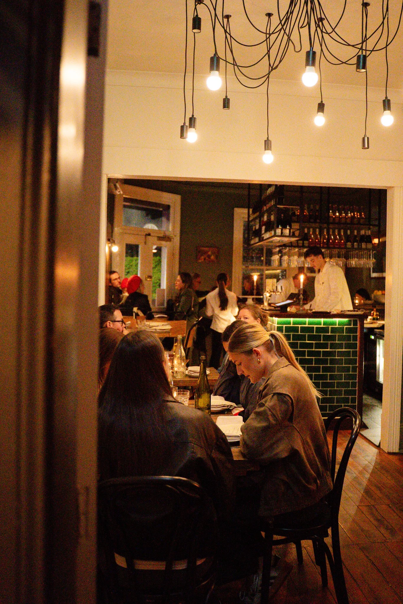Inside a cozy restaurant with hung light bulbs, customers sitting at tables, a bar area with bottles, and staff working behind the counter.