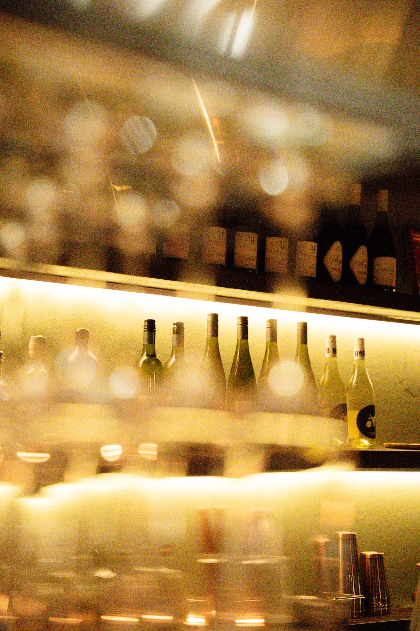 Blurred view of a bar shelf with bottles of alcohol illuminated by warm lighting.