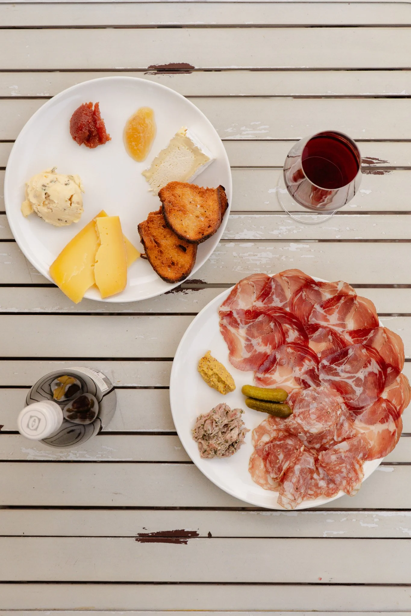 A spread of sliced cured meats, pickles, mustard, and tuna with a plate of cheese, toasted bread, mashed potatoes, fruit preserves, and bread slices, alongside a glass of red wine and a bottle of water on a weathered outdoor table.