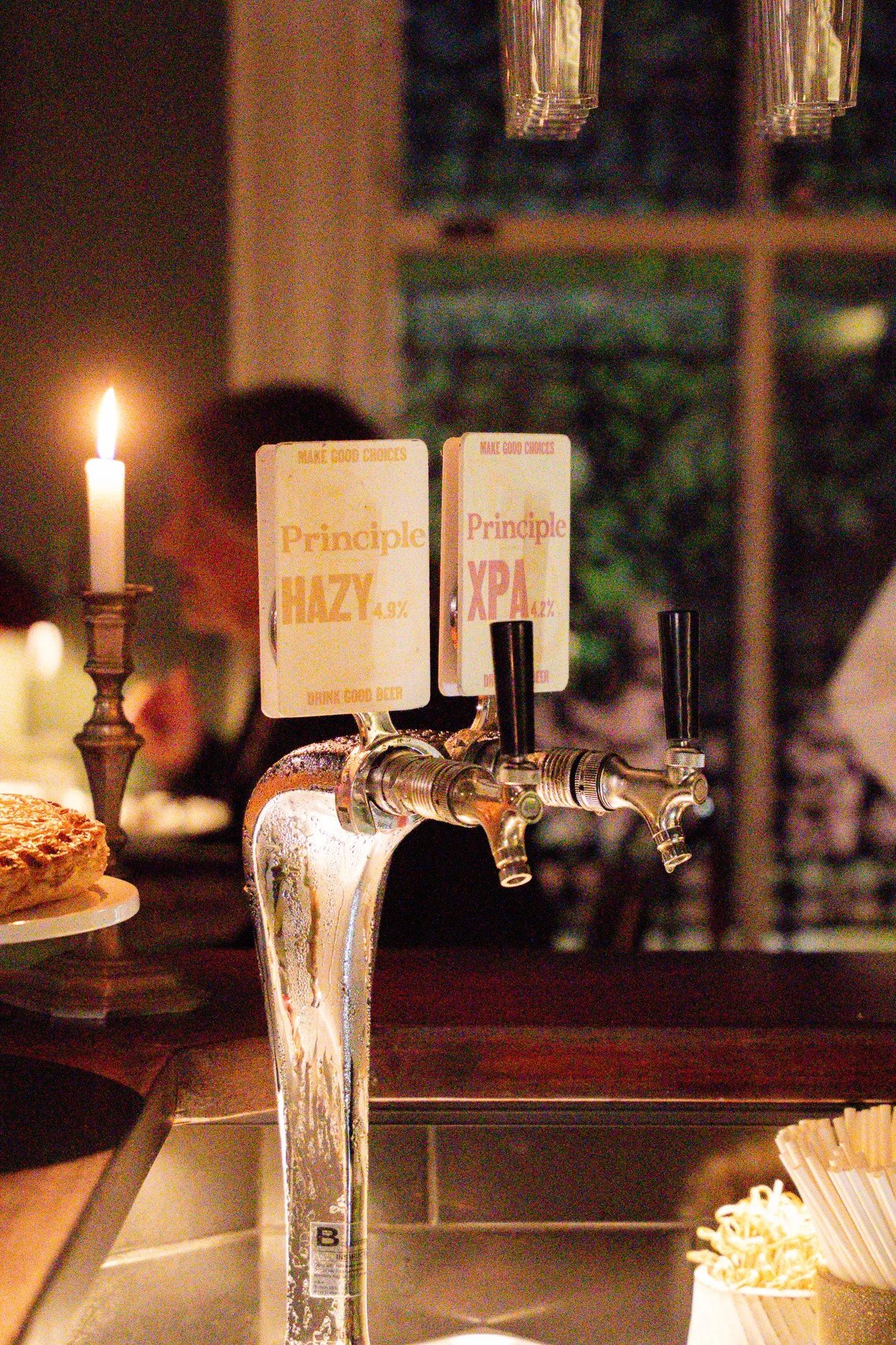 A beer tap with two labels, one showing 'Hazy 4.9%' and the other 'XPA 4.2%', in a bar setting with a candle and blurred background.