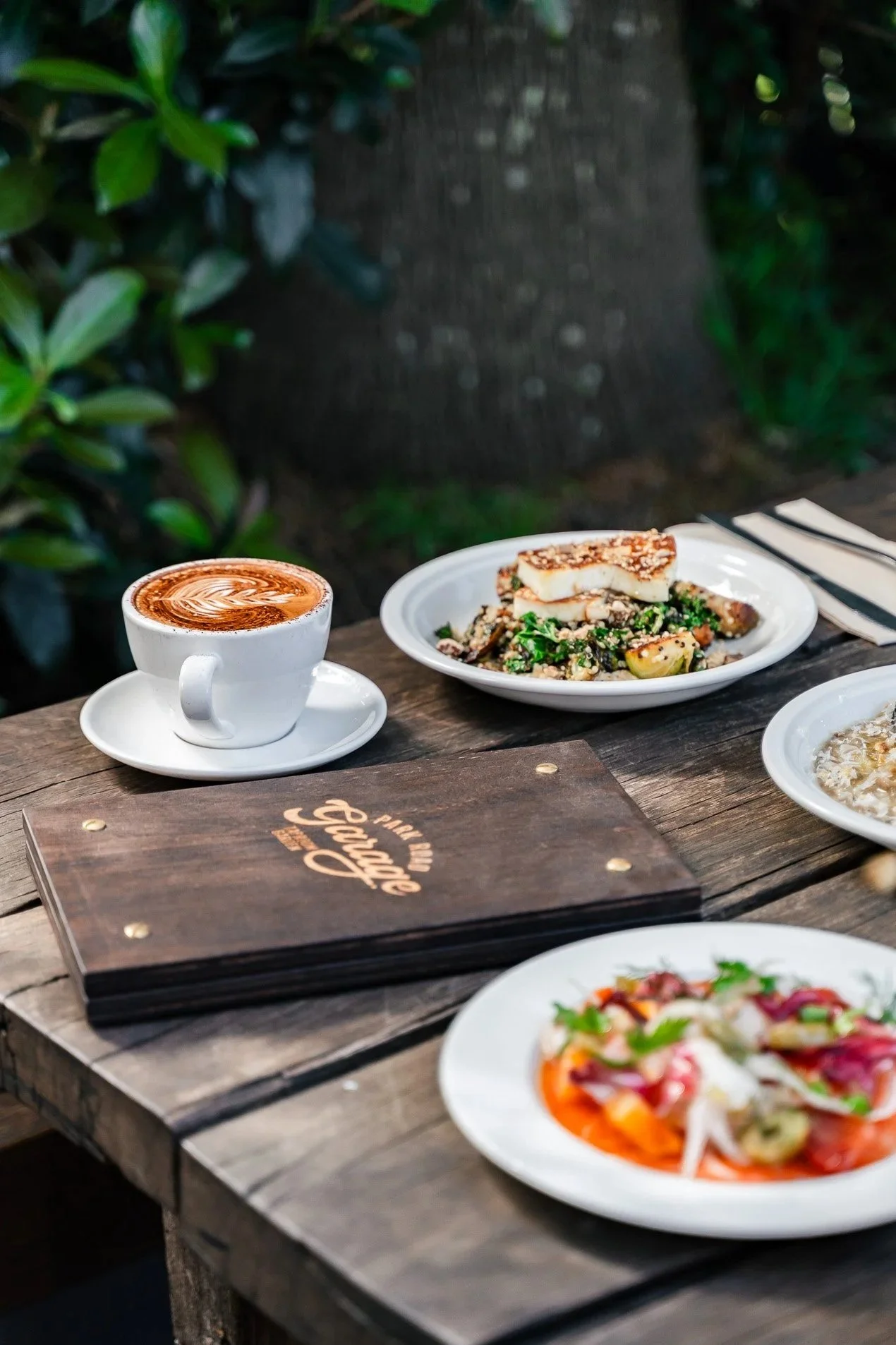 A wooden table with a cup of coffee, a menu, and several plates of food, set outdoors with green foliage in the background.