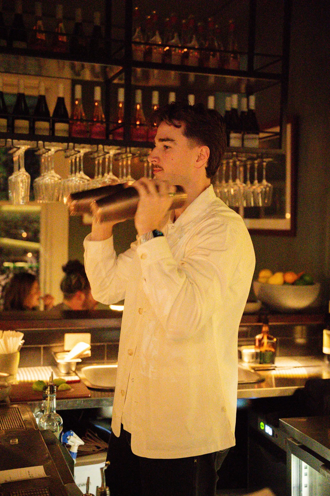 A male bartender in a white jacket shakes a cocktail shaker behind a bar with glasses and bottles visible in the background.
