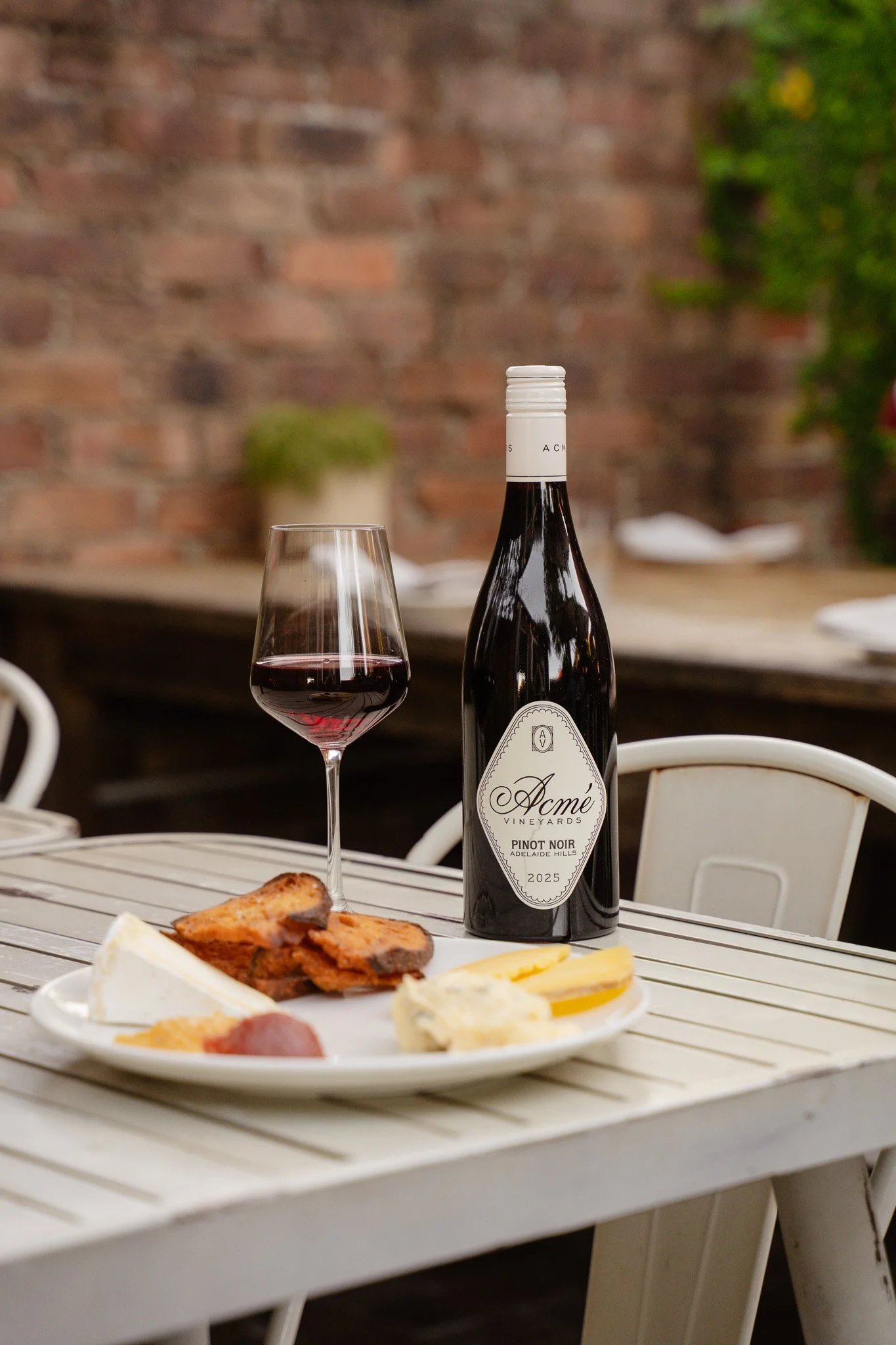 A table set with a glass of red wine, a bottle of Acmé Vineyards Pinot Noir, and a white plate with various cheeses and meats, against a brick wall background.