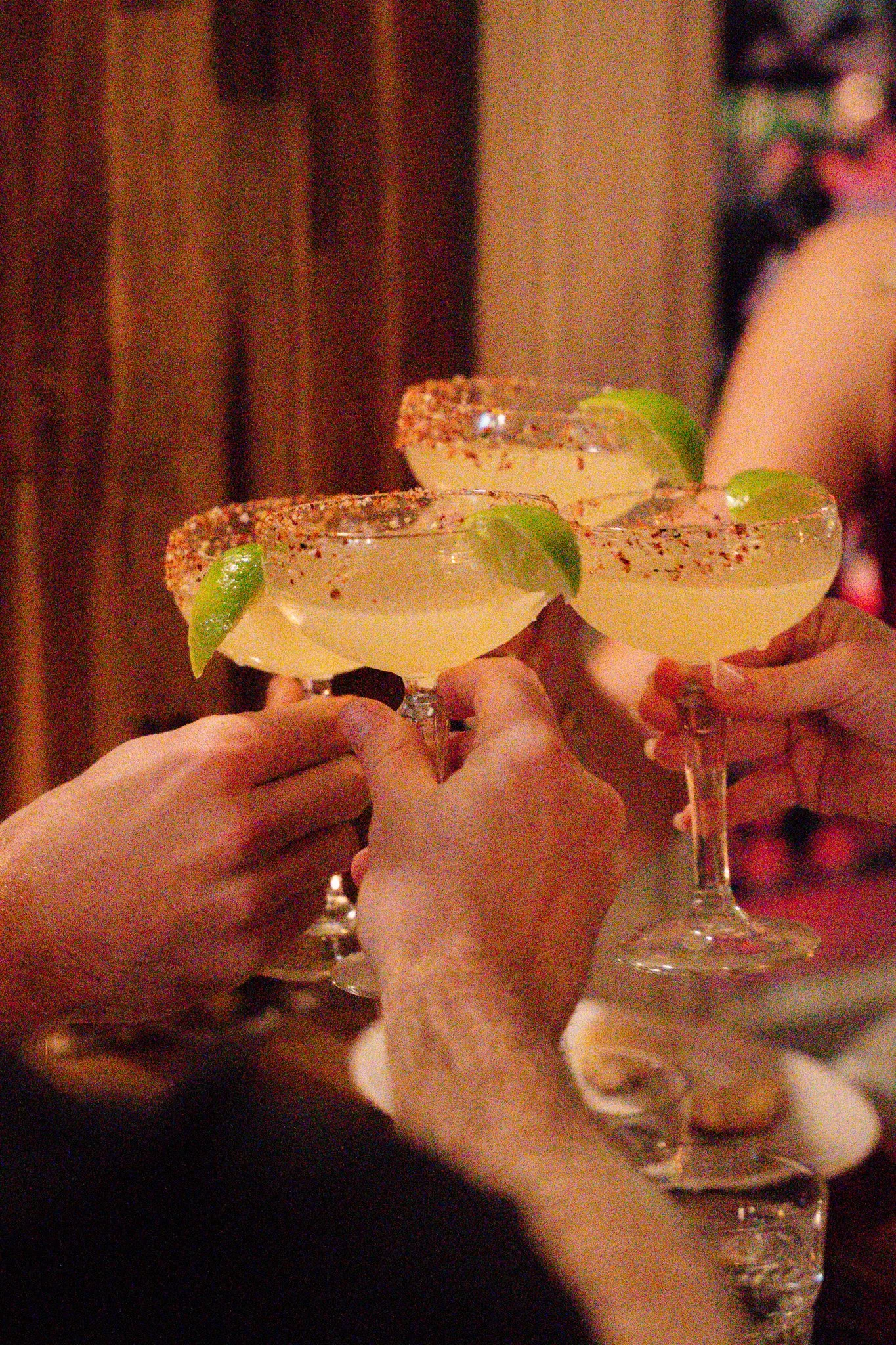 Three people clinking margarita glasses garnished with lime wedges, with salted rims, in a dimly lit setting.