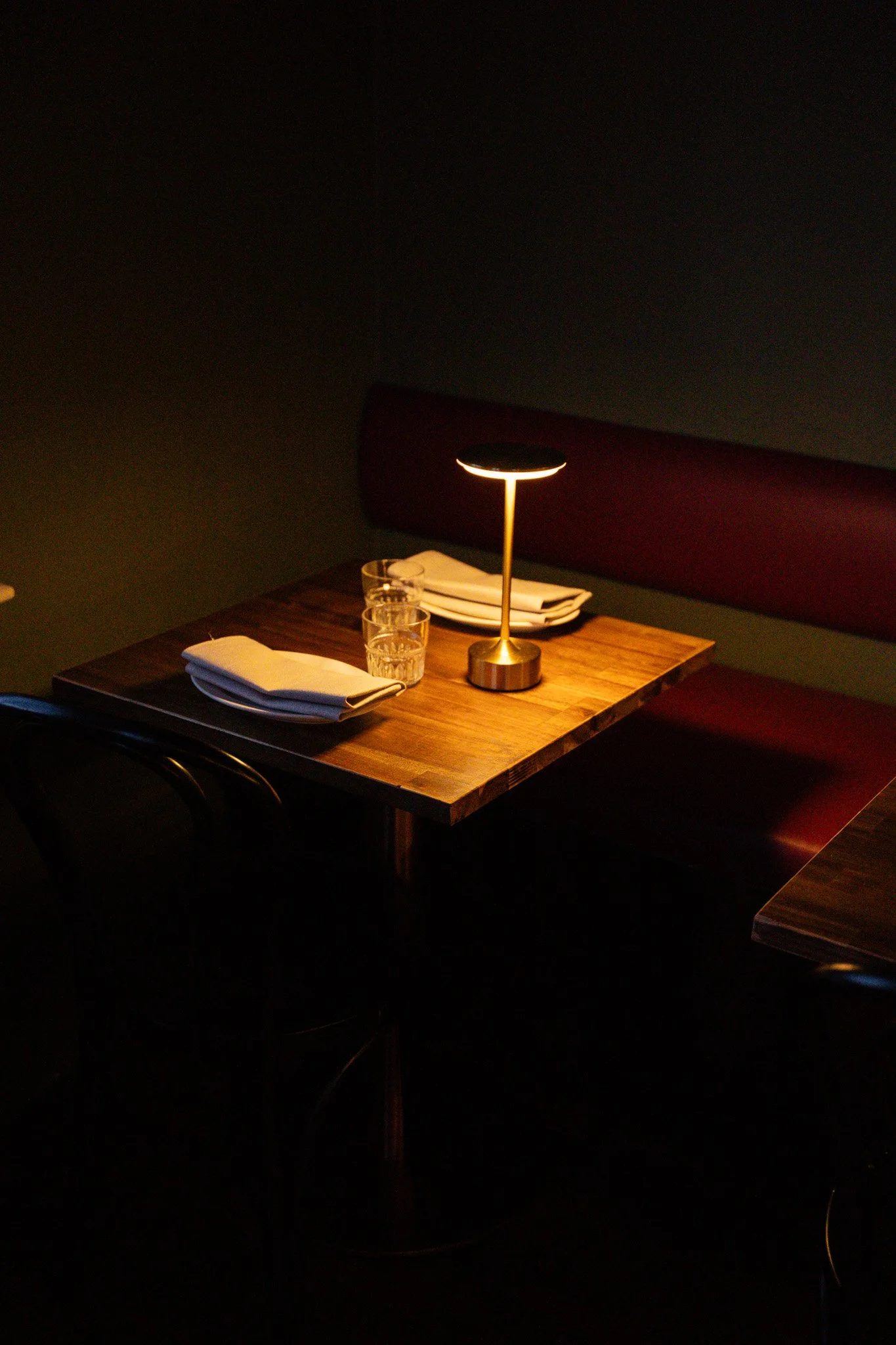 Empty restaurant table with two glasses, folded napkins, and a brass table lamp in a dimly lit setting.