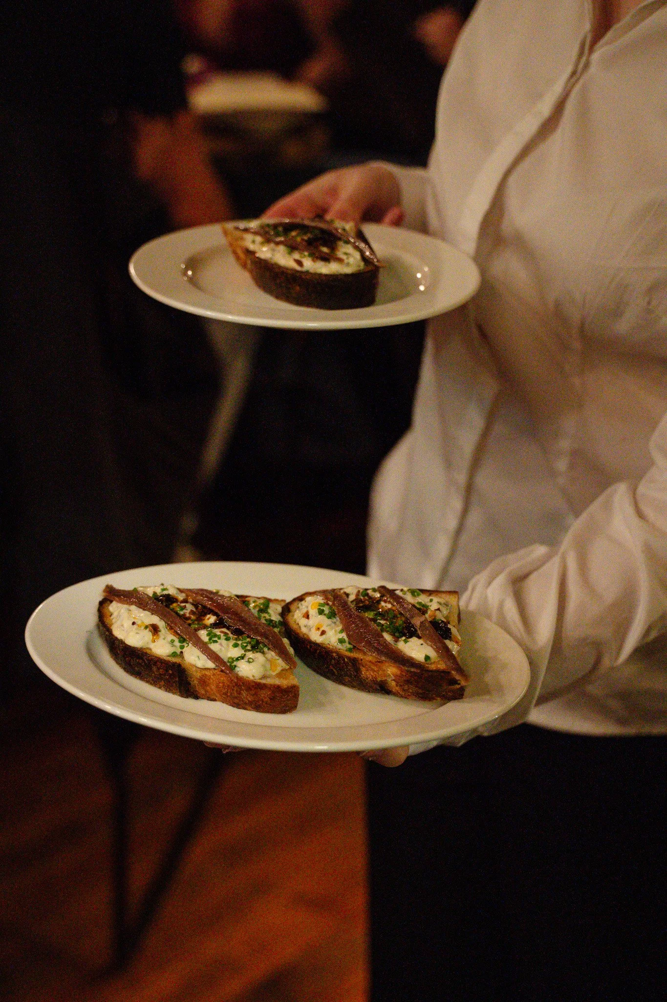 A server holding two plates of food, with the foreground plate featuring two slices of toasted bread topped with cream, chives, and bacon, and the background plate showing a halved baked potato with toppings.