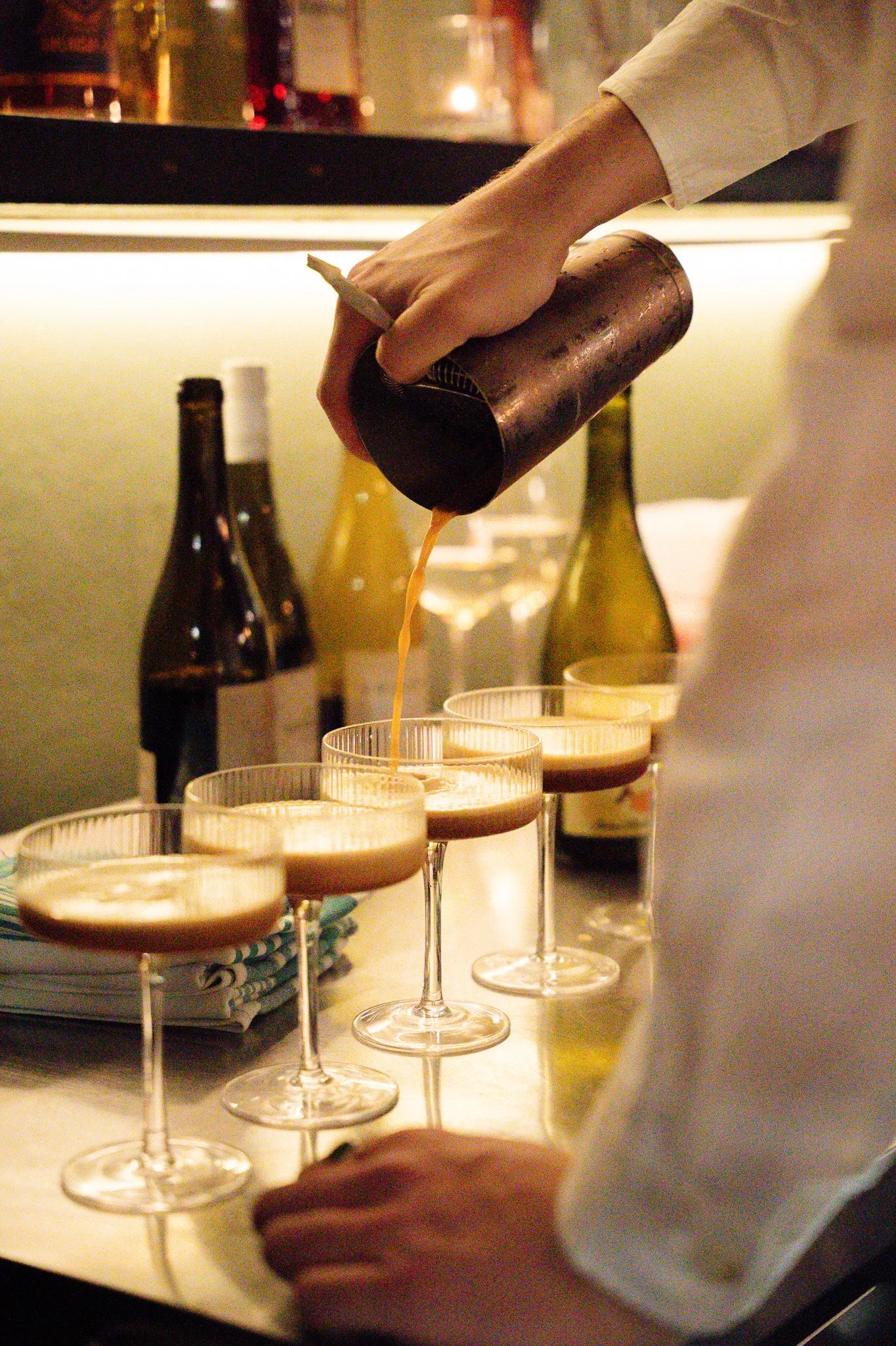 A bartender pours a drink into four glasses with a cocktail shaker in hand, behind a bar with bottles in the background.