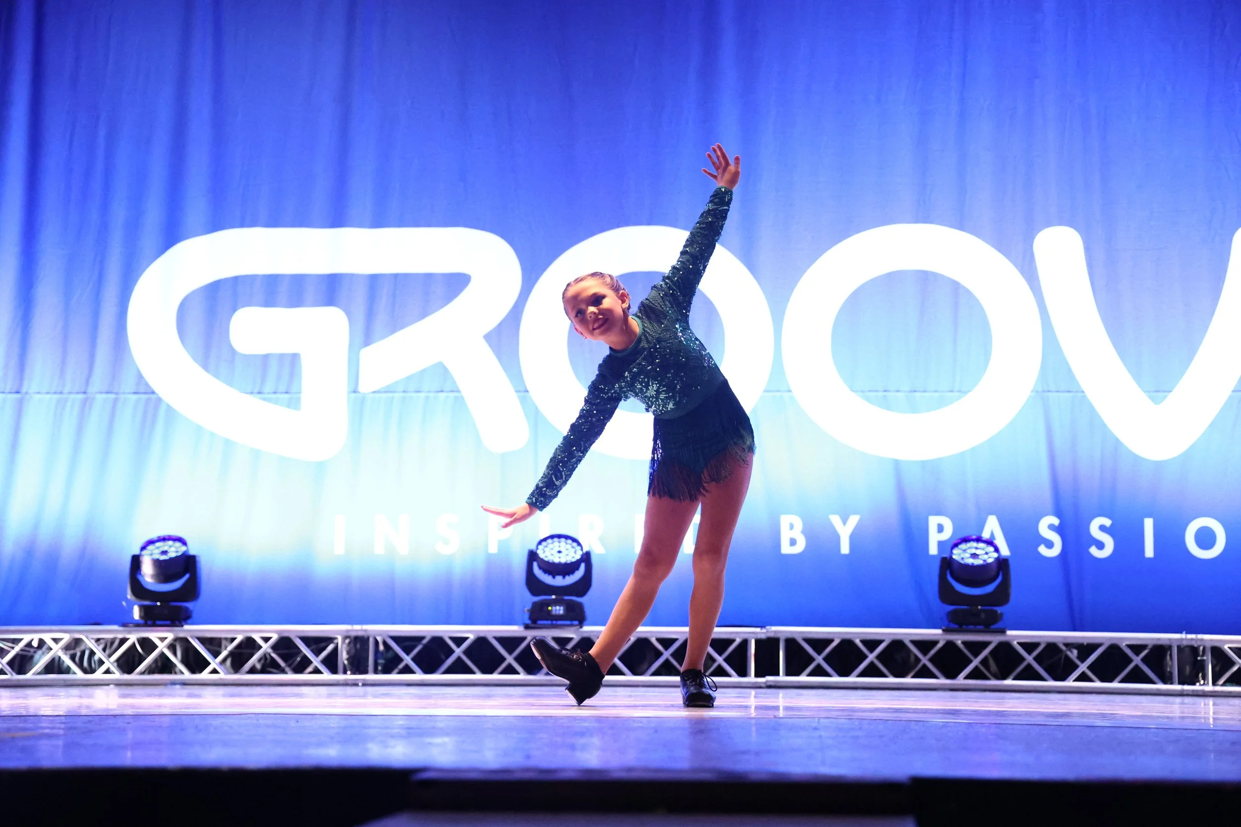 Young girl performing a dance move on stage at an event with a large blue backdrop that reads 'GROOVY' and 'INSPIRATION BY PASSION', wearing a sparkly dark blue top, a fringed skirt, and black shoes.