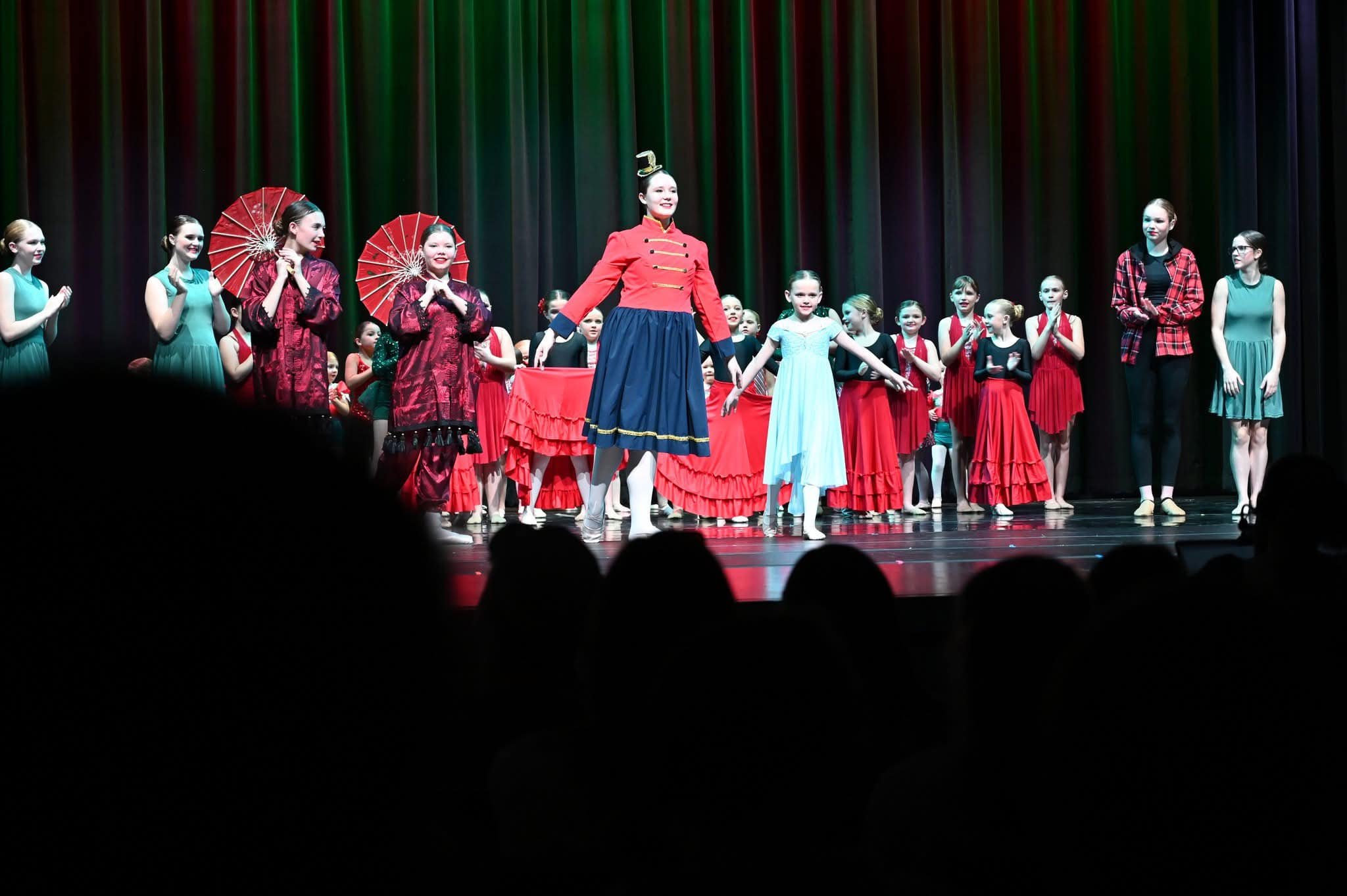 Children and adults on stage after a dance performance, dressed in colorful costumes, with some holding umbrellas, in front of a dark curtain.