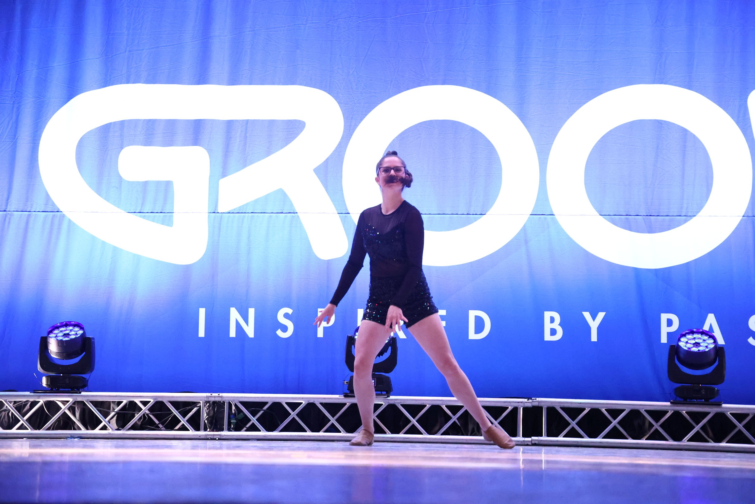 A dancer on stage in front of a large blue backdrop with the Gloo logo, performing a dance move with one hand on her thigh, wearing a black sequin outfit and tan dance shoes.