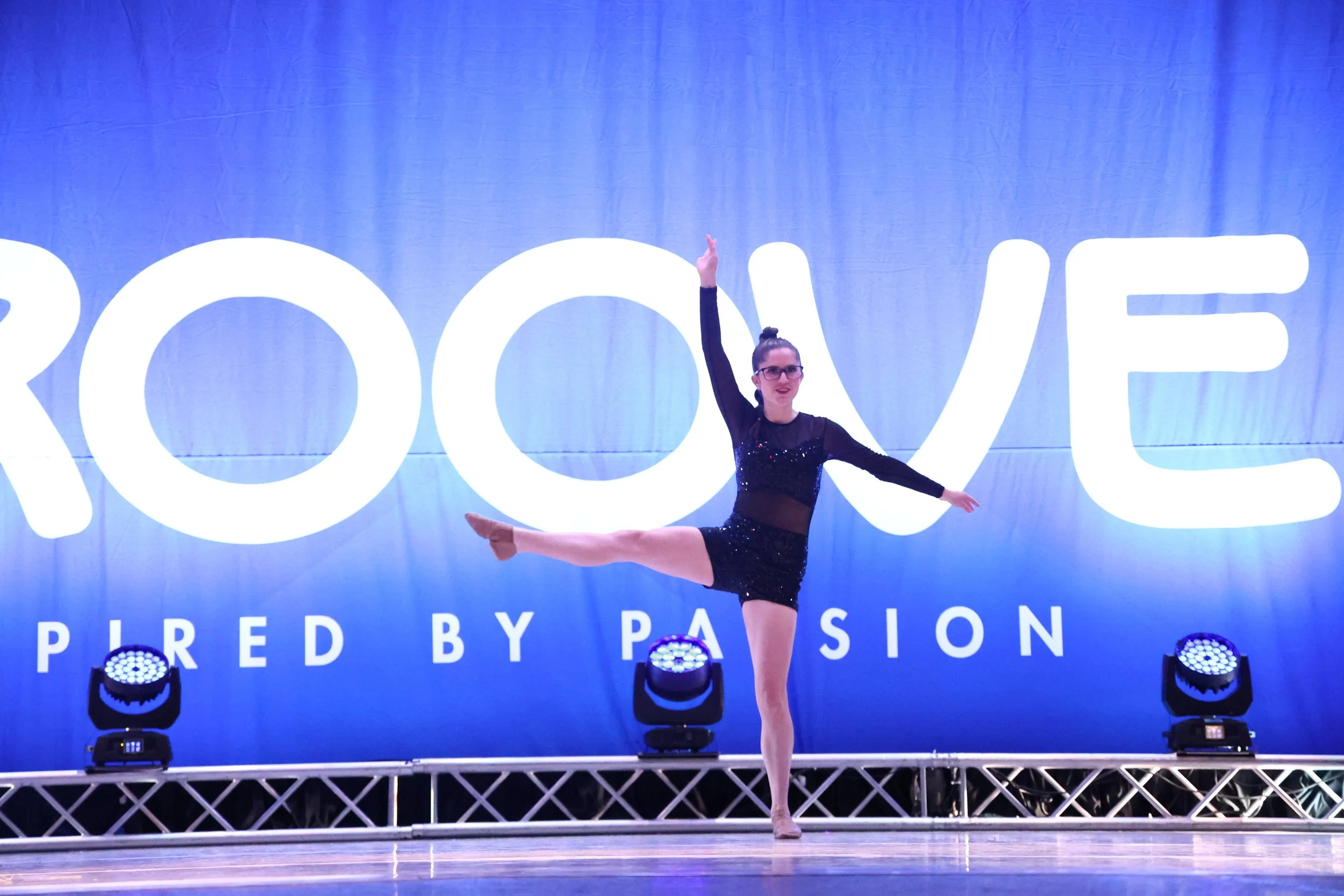 A female dancer performing a pose on stage with a large illuminated 'ROOVE' logo in the background, wearing a black outfit decorated with sequins.