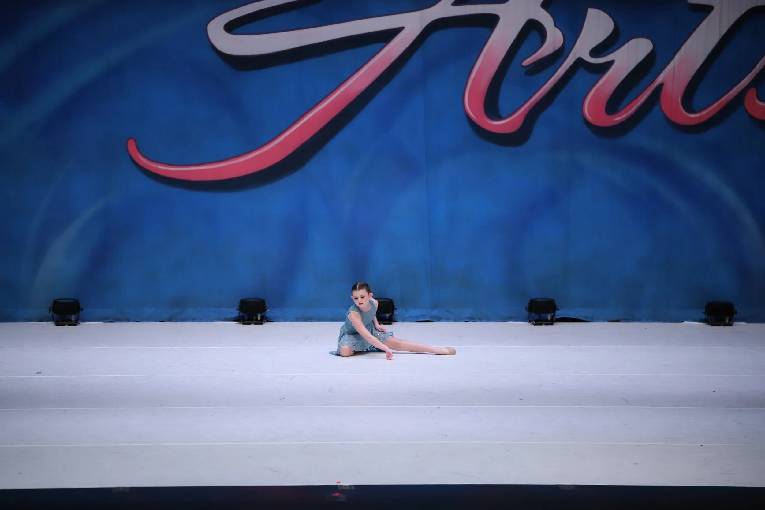 A young girl in a dance costume performing a dance move on stage with a large blue backdrop that has cursive text.