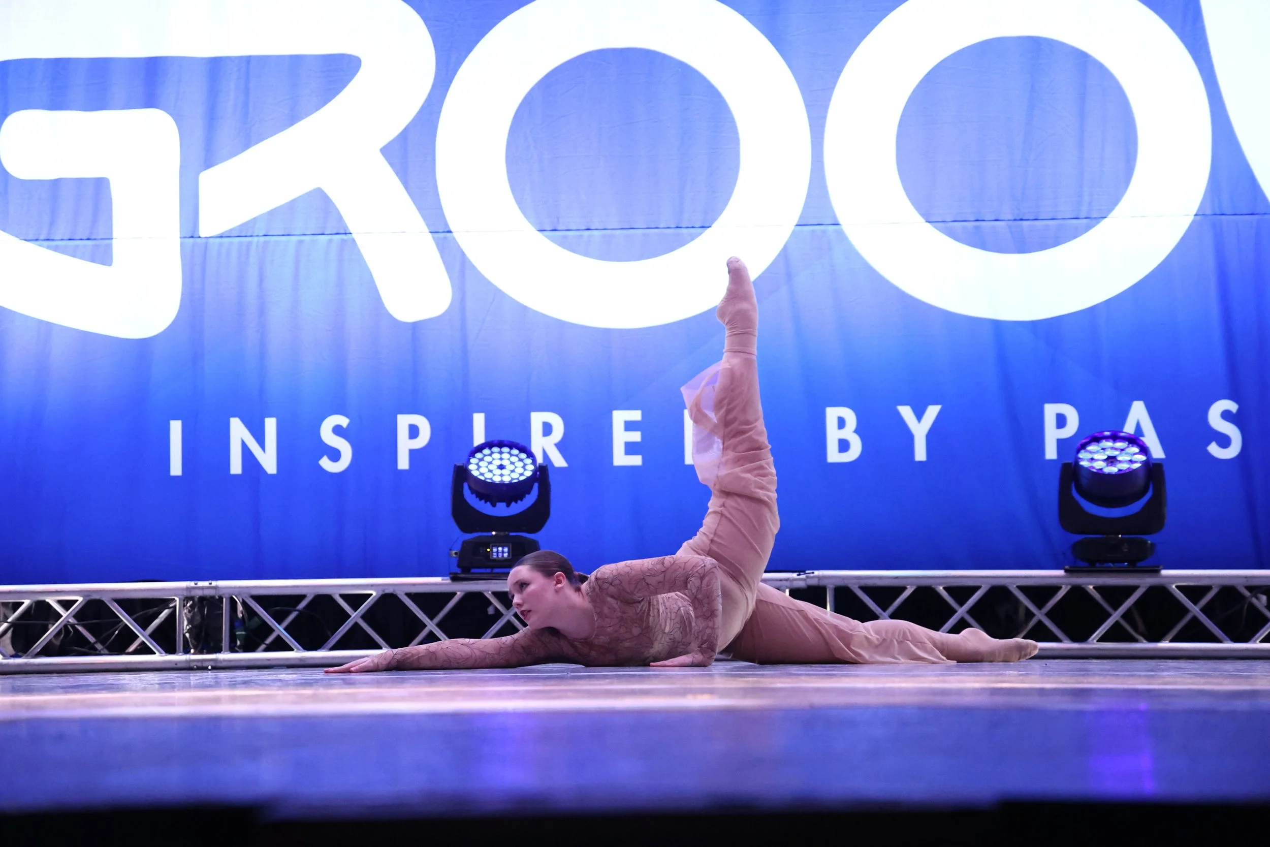 A female gymnast performing a floor routine on stage with a large blue backdrop that reads 'GROOV' and 'INSPIRE BY PAS'. She is lying on her side with one arm extended on the floor and her other leg raised high in the air.