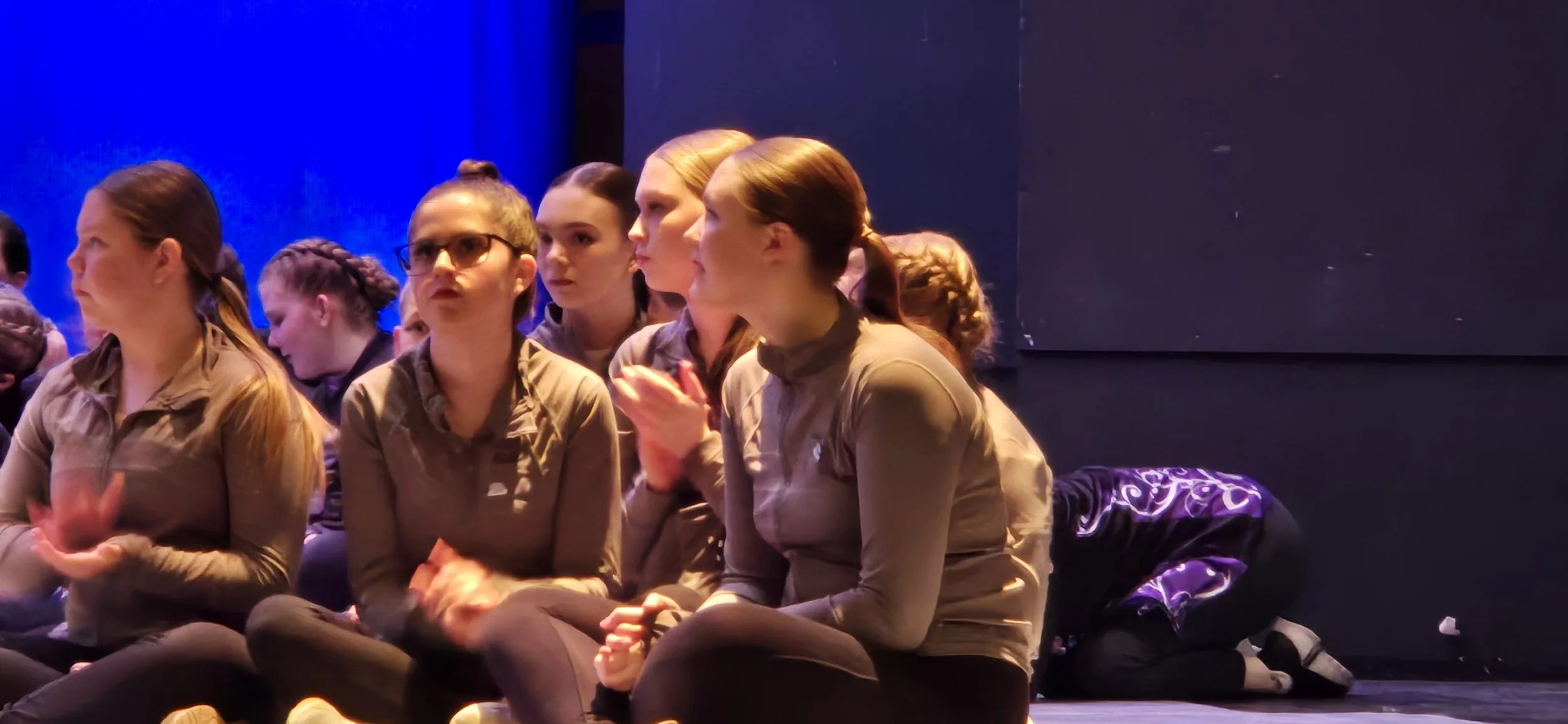 Group of young women sitting on a stage, clapping and watching a performance that appears to be a play or dance. One woman is kneeling in the background.