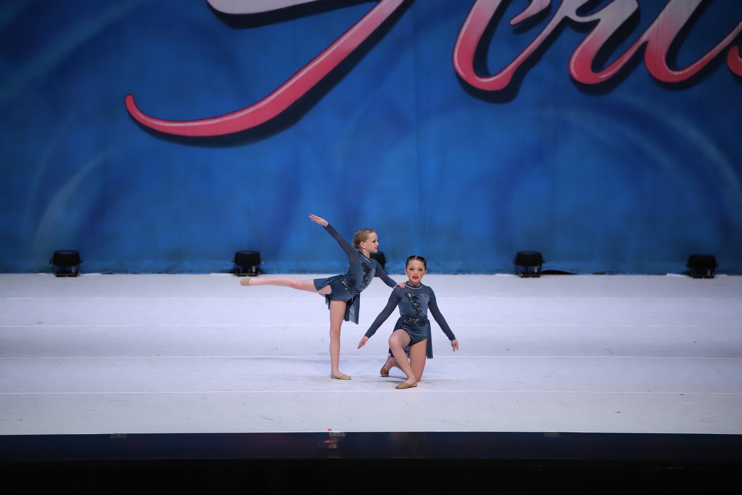 Two young female ballet dancers performing on stage, one stands in arabesque and the other kneels, wearing matching dark costumes with floral embellishments, against a blue backdrop with red script.
