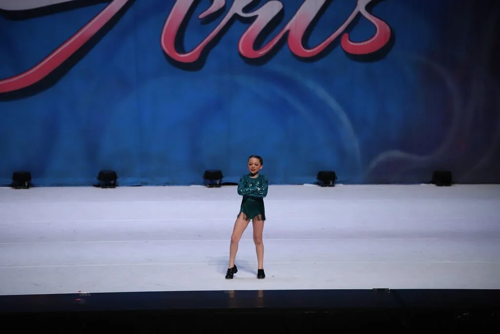 A young girl performing a dance routine on a stage with a blue backdrop and red cursive script.